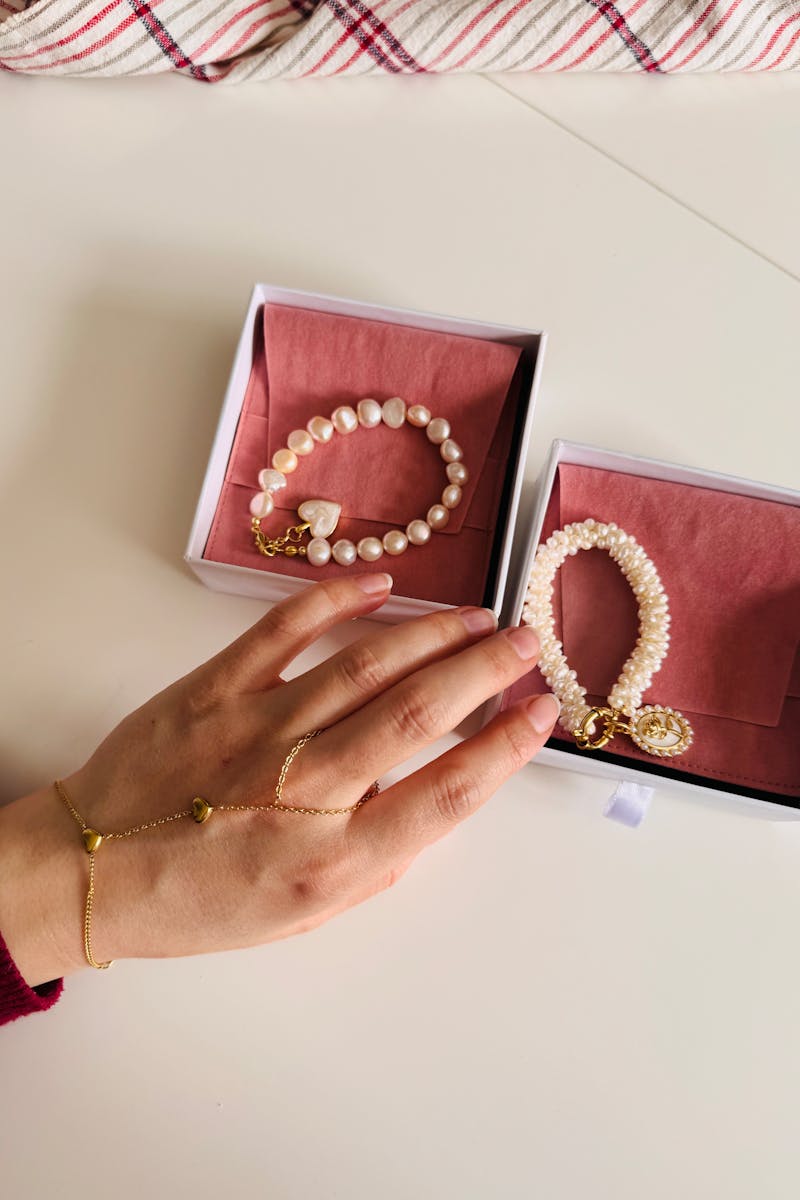 Hand model displaying pearl bracelets with gold accents in open gift boxes.
